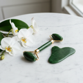 Jade roller and gua sha set on white marble surface with orchid flowers and green leaves